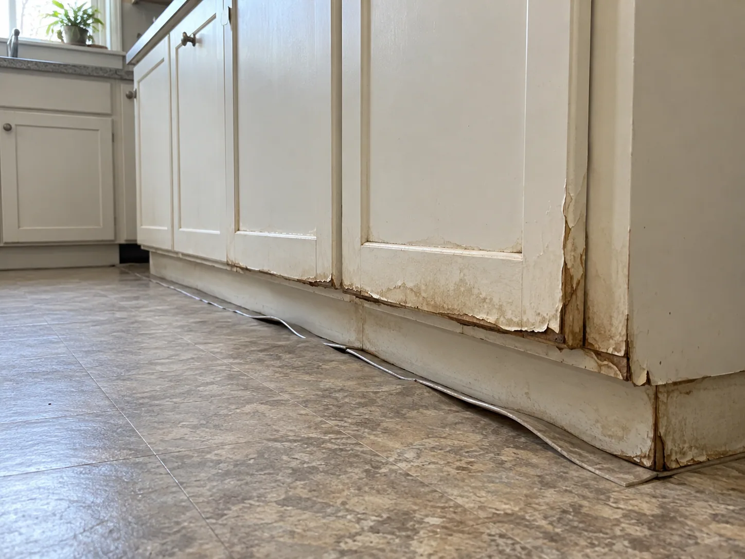 Swollen kitchen cabinet plinth and lifting vinyl flooring from water damage in a Dallas home